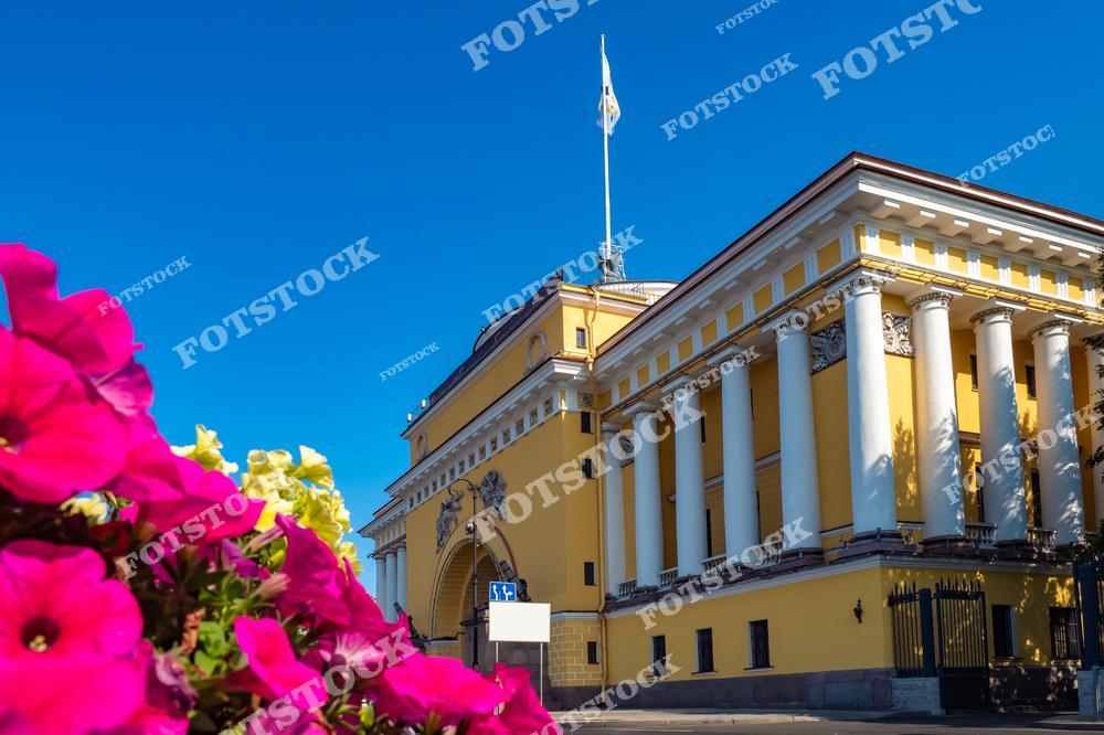 На фотографии изображено здание Адмиралтейства — одна из главных достопримечательностей Санкт-Петербурга. Этот архитектурный памятник с колоннадой и высоким шпилем является символом города. Цветы на переднем плане добавляют ярких красок и подчеркивают летнюю атмосферу. Голубое небо делает снимок особенно выразительным.
